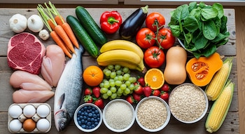 Vista cenital de una mesa de madera rústica con una abundante selección de alimentos frescos que incluyen carne roja, pollo, pescado, huevos, vegetales, frutas y cereales.