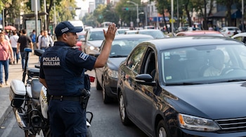 Un oficial de policía con uniforme azul, chaleco y gorra se para junto a una motocicleta policial, con la mano derecha levantada frente a un coche oscuro.