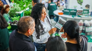Una mujer de cabello oscuro muestra una bolsa de vermicompost a dos personas; detrás se ven plantas, bolsas de compost y botellas