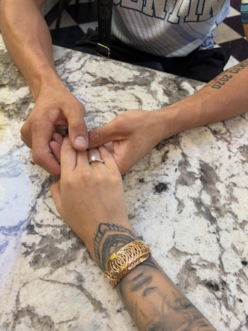 Dos personas entrelazan sus manos sobre una mesa de mármol; una lleva un anillo de compromiso, un brazalete de oro y tatuajes en el brazo