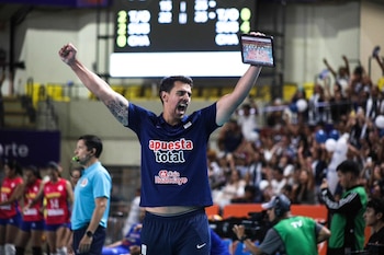 Un entrenador de vóley con camiseta azul y tatuajes en los brazos celebra con los brazos levantados y un tablet en una mano, en una cancha con público al fondo