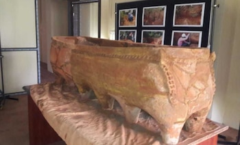 Vasija antigua de terracota con decoraciones y patas, exhibida sobre una mesa. Al fondo, un panel negro con fotos de excavaciones arqueológicas