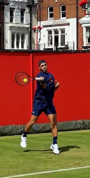 Francisco Cerúndolo bate uma bola de tênis com uma raquete em uma quadra gramada. Ele está vestindo um terno esportivo escuro, um chapéu azul, com uma parede vermelha e um prédio ao fundo