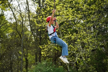 Una joven con casco rojo y arnés azul se desliza por una tirolesa en un bosque, sus brazos levantados, con árboles verdes de fondo