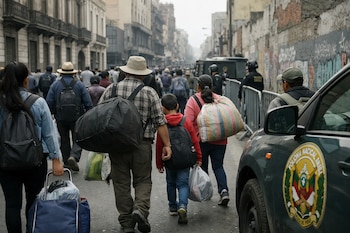 Una multitud de personas camina por una calle urbana. Llevan mochilas, bolsas y un carrito. A la derecha, el costado de un vehículo policial con un emblema.