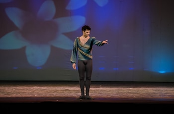Hombre de cabello oscuro con traje de ballet azul verdoso y mallas negras, con un brazo extendido, en un escenario de madera. Proyección azul con forma de flor en el fondo