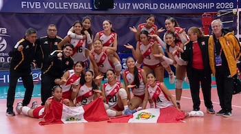 Equipo femenino de voleibol de Perú y su cuerpo técnico posan en cancha. Varias jugadoras sostienen banderas peruanas, con letrero de campeonato de fondo.