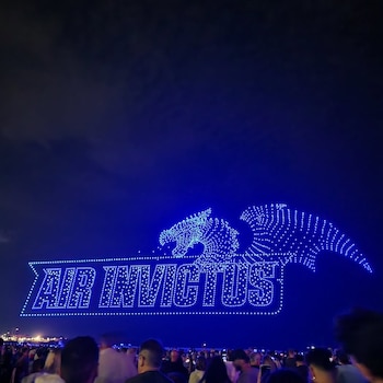 Cielo nocturno azul oscuro con el texto luminoso 'AIR INVICTUS' y una figura de dragón, ambos formados por luces azules. Una multitud mira el espectáculo