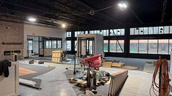 Vista interior de un espacio comercial en construcción con techo oscuro, paredes de ladrillo visto, grandes ventanales y herramientas en el suelo