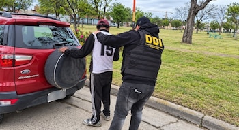 Un hombre apoya las manos en un vehículo rojo mientras un oficial con chaleco que lleva la inscripción DDI lo sujeta por el hombro