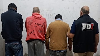 Cuatro hombres de espaldas, tres con ropa casual y uno con chaleco negro que dice PDI. Están frente a una pared blanca con inscripciones