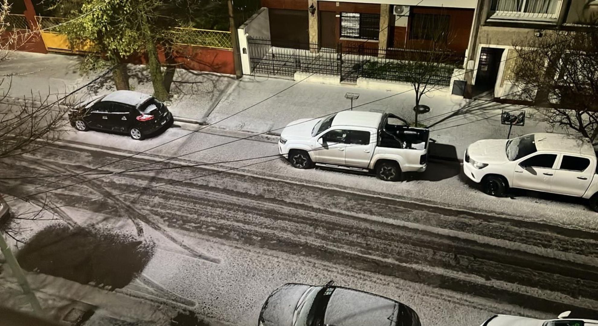 Varios vehículos permanecen estacionados en una calle residencial cuya calzada y aceras se encuentran cubiertas por una capa de nieve durante la noche.