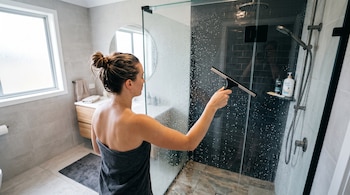 Una persona vista de espaldas con una toalla pasa un secador de goma sobre la mampara de vidrio con agua dentro de un baño.