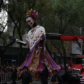Figura gigante de esqueleto con flores y vestido tradicional, guiada por personas en una calle con público y un camión rojo