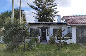 Vista externa de uma casa branca com telhado de palha e jardim com grama, arbustos e duas araucárias, a grande nos fundos e a pequena na frente.