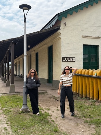 Dos mujeres con gafas de sol posan junto a un farol y frente a un edificio blanco con el cartel Lules