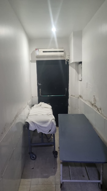 Pasillo de hospital blanco con dos camillas, una con sábanas blancas y otra azul, frente a una puerta oscura. Un aire acondicionado en la parte superior