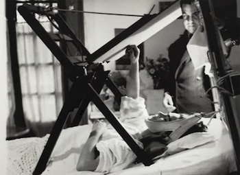 Una mujer recostada en una cama de sábanas blancas utiliza un atril de madera para pintar mientras un hombre de traje la observa