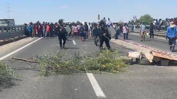 Policías con escudos y cascos frente a un grupo de manifestantes en una carretera bloqueada con ramas y cartones, bajo un cielo claro