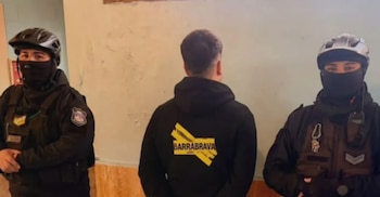 Un hombre visto de espaldas con un buzo negro permanece custodiado por dos agentes de policía que llevan cascos y tapabocas