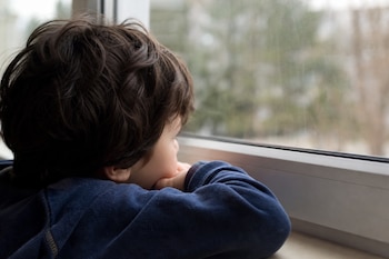 Un niño de cabello oscuro apoyado sobre sus manos mientras mira a través de una ventana.