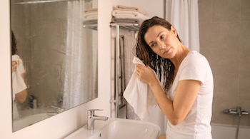 Mujer con camiseta blanca seca su cabello castaño y húmedo con una funda de almohada blanca frente al espejo de un baño.