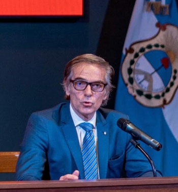 Retrato del juez Gustavo Hornos, un hombre con traje azul y gafas, sentado detrás de un podio con un micrófono y una bandera