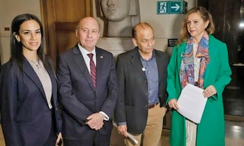Cuatro personas posan de pie en un interior: dos mujeres y dos hombres. Al fondo se observa una escultura, una pared de madera y un cartel de salida