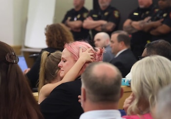 Primer plano de una mujer con cabello rosa, con lágrimas en los ojos, abrazando a una niña con coletas en un tribunal; otras personas borrosas de fondo
