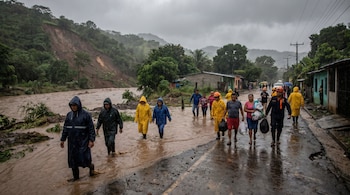 Personas caminan por una calle inundada y un río crecido, con un gran deslave visible en la colina al fondo, bajo un cielo gris y lluvioso.