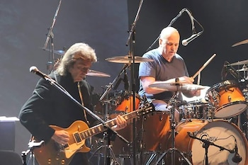 Dos músicos en el escenario. Un guitarrista toca una guitarra eléctrica dorada mientras un baterista toca la batería, que incluye bombo y caja, con micrófonos alrededor