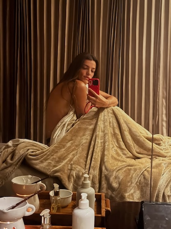 Calu Rivero, envuelta en una manta dorada, se toma una selfie frente a un espejo, con productos de spa en primer plano y cortinas marrones al fondo