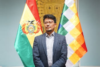 Fernando Aramayo, un hombre con gafas y barba, lleva un traje azul sobre una camisa a cuadros, de pie ante las banderas de Bolivia y la Wiphala