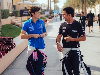 Dos hombres caminan por un sendero con palmeras a los lados; uno viste camiseta azul y pantalón negro con detalles rosas, el otro camiseta negra y pantalón a juego