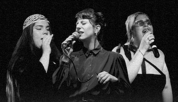 Fotografía en blanco y negro de tres mujeres cantando en un escenario, cada una con un micrófono en la mano