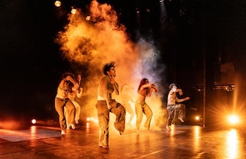 Un grupo de bailarines realiza movimientos sobre un escenario iluminado con luces cálidas y humo denso en el centro