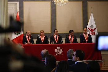 Cinco funcionarios, cuatro hombres y una mujer, con vestimenta formal y bandas, sentados en una mesa roja con el logo JNE, frente a una audiencia. Banderas peruana e INE visibles