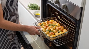 Una persona con delantal introduce una fuente de vidrio con papas pequeñas, ramas de romero y especias en un horno abierto.