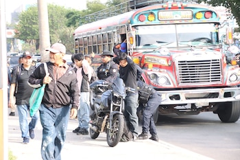 Agentes de policía inspeccionan un autobús de pasajeros y una motocicleta en una calle con civiles cerca. El autobús es rojo y azul, la motocicleta es negra