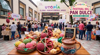 Cestas con pan dulce tradicional y una taza con chocolate caliente sobre una mesa de madera, con puestos y asistentes en un patio al fondo.