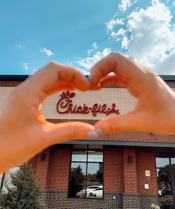 Manos de una persona forman un corazón frente a la fachada de ladrillo de un restaurante Chick-fil-A, con su letrero rojo y ventanas
