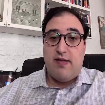 Hombre con gafas y camisa a rayas sentado frente a una pared blanca con estanterías, copas, libros y un cuadro
