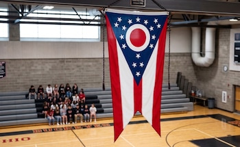 Bandera de Ohio cuelga en el centro de un gimnasio. Al fondo, estudiantes sentados en gradas grises sobre una cancha de baloncesto de madera
