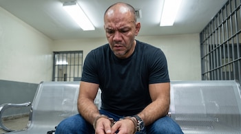 Hombre calvo con camiseta oscura y jeans sentado en un banco metálico. Lleva esposas en las muñecas y un reloj. Al fondo se observan rejas y puertas.