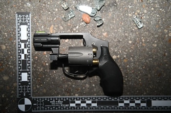 Pistola revólver negra y gris con dos cartuchos, dos reglas de medición forenses blancas y negras, fragmentos de vidrio y un objeto marrón sobre suelo de asfalto