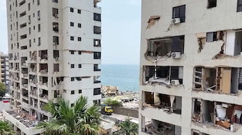 Dos edificios con fachadas dañadas, ventanas rotas, balcones destruidos, escombros, palmeras, un camión amarillo y el mar de fondo