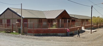 Un edificio escolar con paredes de ladrillo, cerco con grafitis y techos de chapa, con personas paradas en una calle de tierra