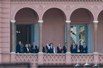 Grupo de personas en un balcón con arcos y columnas de un edificio rosado; el hombre central en chaleco azul levanta la mano, otros aplauden