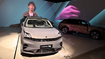 Dos vehículos Lynk&Co en un showroom: un Lynk&Co 06 gris al frente y un modelo marrón detrás, con proyecciones de un hombre y una mano al fondo
