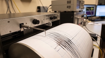 Un sismógrafo registra datos en un rollo de papel cuadriculado. Se ven equipos electrónicos, cables y un monitor de computadora al fondo.